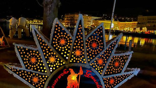 A star-shaped Glöckler cap with the illuminated inscription ‘Feuerwehr Gmunden’ (Gmunden Fire Brigade) and bright patterns against a night-time cityscape, whose bright lights are reflected in the water.