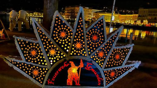 Eine sternförmige Glöcklerkappe mit der leuchtenden Aufschrift "Feuerwehr Gmunden" und leuchtenden Muster vor einer nächtlichen Stadtlandschaft, deren helle Lichter sich im Wasser spiegeln.