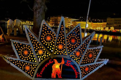 A star-shaped Glöckler cap with the illuminated inscription ‘Feuerwehr Gmunden’ (Gmunden Fire Brigade) and bright patterns against a night-time cityscape, whose bright lights are reflected in the water.