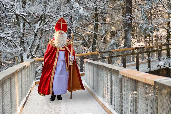 Eine als Nikolaus verkleidete Person geht über eine verschneiten Holzweg in einem Wald, trägt ein rotes Gewand und eine rote Mütze und hält einen Stab in der Hand.