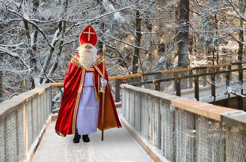 A person dressed as St. Nicholas walks along a snow-covered wooden path in a forest, wearing a red robe and a red cap and holding a staff in his hand.