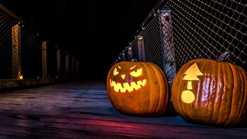 Two glowing carved pumpkins, one scary and one with a tree, sit on a dark wooden walkway at night.