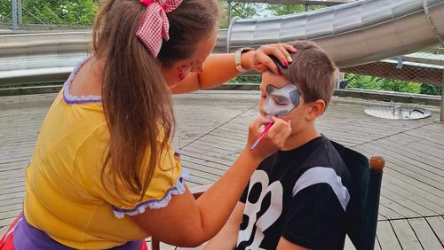 Woman face paints a boy's face with a gray design; a large metal slide is in the background.