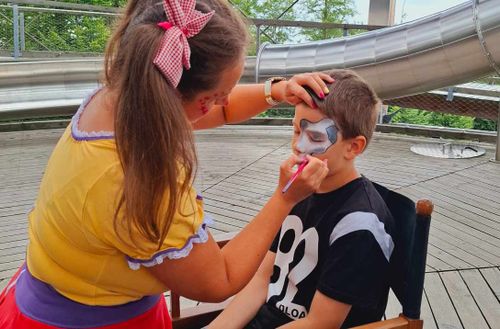 Woman face paints a boy's face with a gray design; a large metal slide is in the background.