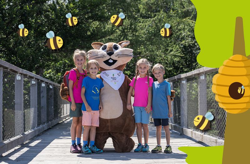 Kinder und ein Eichhörnchen-Maskottchen stehen auf einer Holzbrücke, umgeben von fliegenden Comic-Bienen und einem Baum mit Bienenstock.
