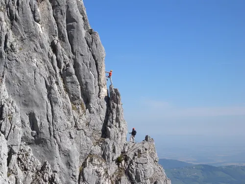 Zwei Personen mit Helm und Gurt sichern sich beim Klettern an einer steilen Felswand mit weitem Ausblick ins Tal.