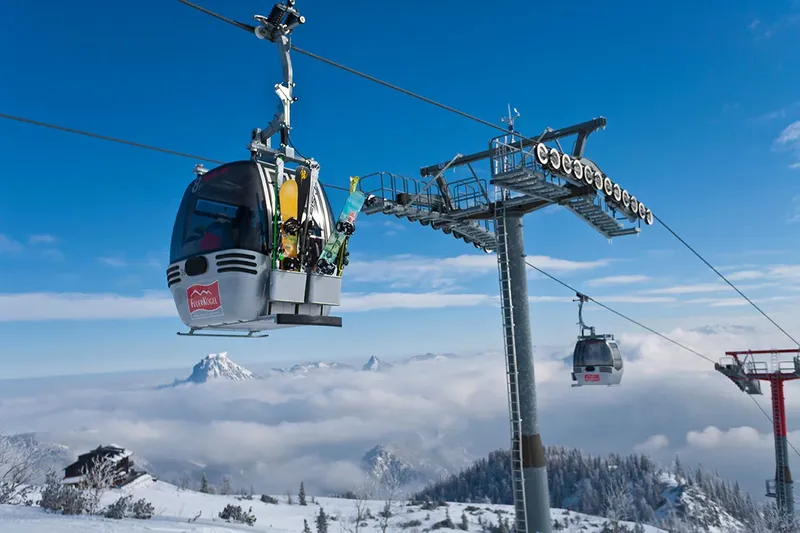 Eine Gondel mit Skiern außen gleitet bei strahlend blauem Himmel über das verschneite Feuerkogel-Plateau und ein Meer aus Wolken.