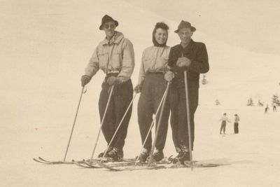 Drei Personen in nostalgischer Skibekleidung posieren mit Skiern auf verschneitem Hang, hinten fahren weitere bei klarem Himmel – Feuerkogel.