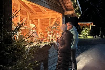 Gemütlicher Weihnachtsmarktstand aus Holz bei Nacht, mit Lichterketten, Schnee und warmem Licht, das Kunden und eine Verkäuferin beleuchtet.