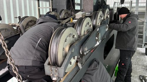Drei Arbeiter in Winterkleidung warten in einer verschneiten Umgebung ein großes Seilbahnsystem mit Seilen.
