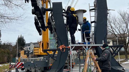 Ein Kran hebt eine große Konstruktion an, während Arbeiter sie auf einem Gerüst zusammenbauen. Auf dem Boden liegen Sonnenkollektoren.