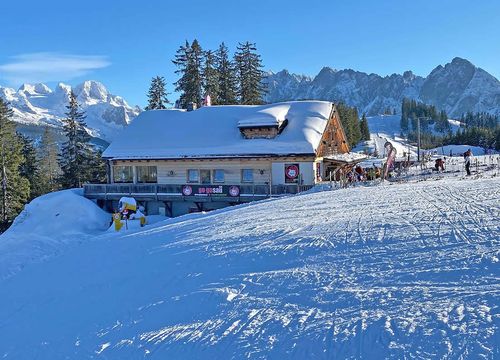 Snow-covered ski hut on a sunny mountainside, surrounded by trees and distant peaks under a clear blue sky. The Dachstein glacier in the background.