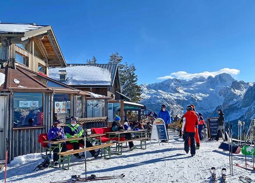 Menschen, die an Tischen im Freien bei einer verschneiten Berghütte sitzen, mit Skiern in der Nähe und einem malerischen Blick auf schneebedeckten Dachstein Gletscher unter einem klaren blauen Himmel.