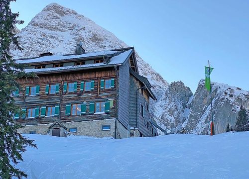 Eine rustikale Berghütte aus Holz und Stein liegt an einem verschneiten Hang, umgeben von hohen Gipfeln und unter einem strahlend blauen Himmel.