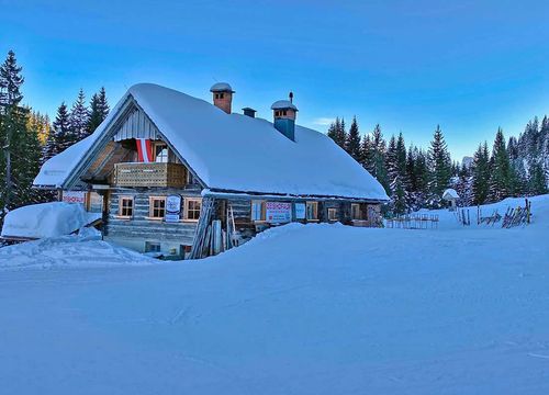 Eine schneebedeckte Holzhütte mit steilem Dach, umgeben von Bäumen unter einem klaren blauen Himmel, in einer Winterlandschaft.