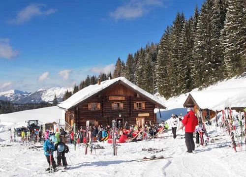 Skifahrer versammeln sich vor einer Holzhütte in einer verschneiten Berglandschaft, umgeben von Bäumen und blauem Himmel.