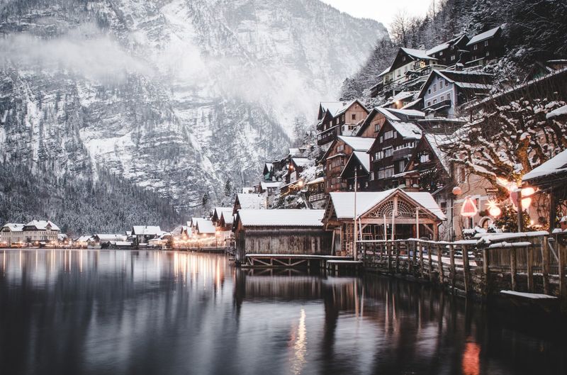 Ein verschneites Dorf an einem ruhigen See, mit Holzhäusern an einem Berghang und nebligen Gipfeln im Hintergrund, beleuchtet von warmen Lichtern.