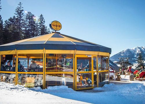 A yellow and blue octagonal umbrella bar on a snowy mountain, surrounded by trees and red deckchairs, with a view of the mountains in the background.