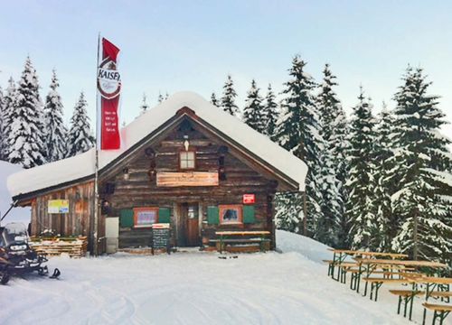 A snow-covered wooden hut with a red flag, surrounded by snow-covered trees and benches outside. A skidoo is parked nearby.