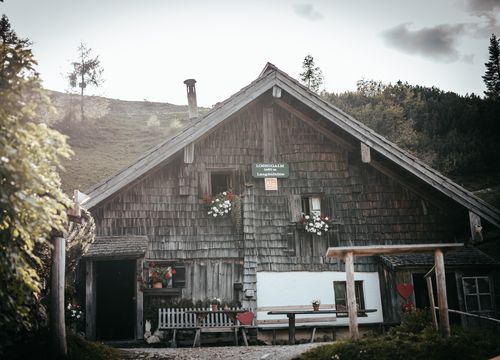Rustikale Holzhütte mit roten Herzdekorationen, Sitzgelegenheiten im Freien und Bergkulisse. Heller, sonniger Tag mit viel Grün rund um die Szene.