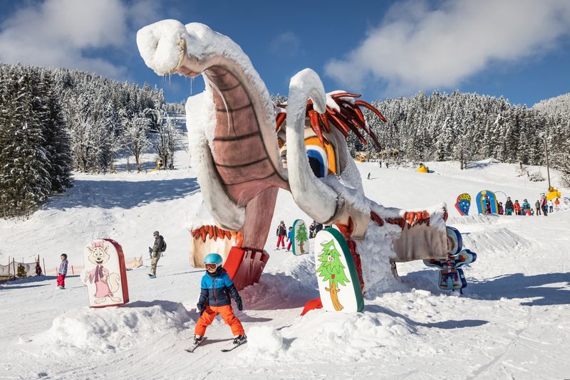 Kind beim Skifahren durch eine große, farbenfrohe Schneeskulptur, die die Form eines Mammuts hat. Umgeben von schneebedeckten Bäumen, bunten Schildern und anderen Wintersportlern.