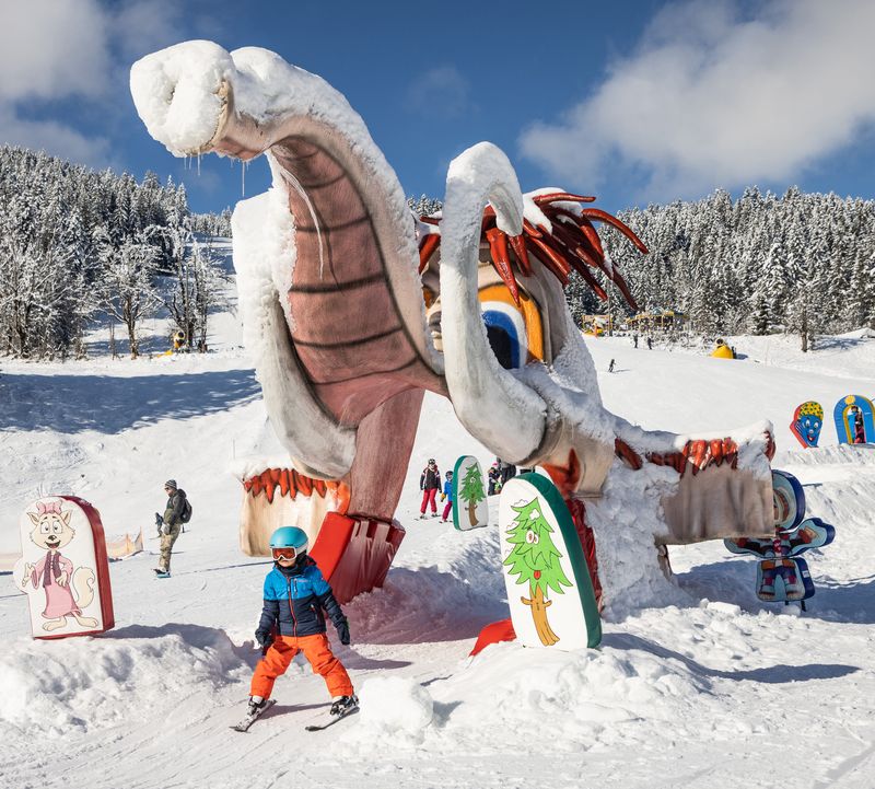 Kind beim Skifahren durch eine große, farbenfrohe Schneeskulptur, die die Form eines Mammuts hat. Umgeben von schneebedeckten Bäumen, bunten Schildern und anderen Wintersportlern.