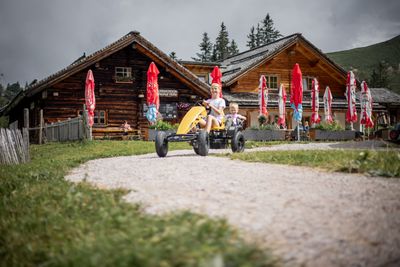 Ein rustikales Holzchalet mit Sitzgelegenheiten im Freien, Kinder die im Vordergrund mit GoKarts fahren, umgeben von grünem Gras und weißen Wildblumen unter einem teilweise bewölkten blauen Himmel.