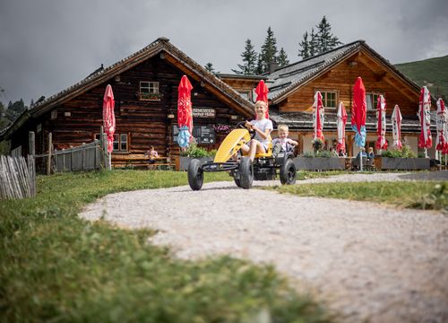 Ein rustikales Holzchalet mit Sitzgelegenheiten im Freien, Kinder, die im Vordergrund GoKart fahren, umgeben von grünem Gras und weißen Wildblumen unter einem teilweise bewölkten blauen Himmel.