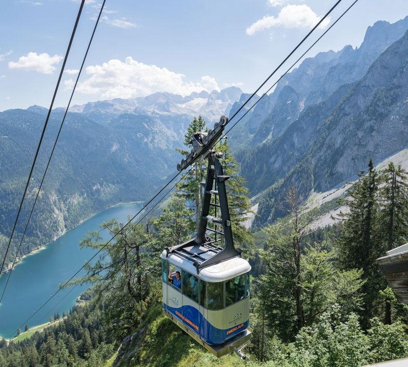 Eine Gondel bei der Bergfahrt, aus dem offenen Fenster winkt eine Person. Rundherum bewaldete Felsformationen, der türkisgrüne Gosausee und der schneebedeckte Dachstein Gletscher.