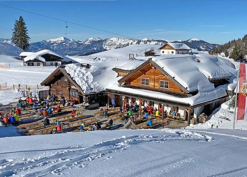 Verschneite Berghütte mit Sitzgelegenheiten im Freien, umgeben von Skifahrern und schneebedeckten Gipfeln unter einem strahlend blauen Himmel.