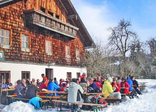 People sitting at outdoor tables on wooden benches and tables, next to a rustic wooden building with a balcony, under a clear blue sky.