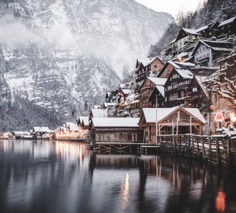 A snowy village on a quiet lake, with wooden houses on a hill and misty mountains in the background, creating a tranquil winter scene.