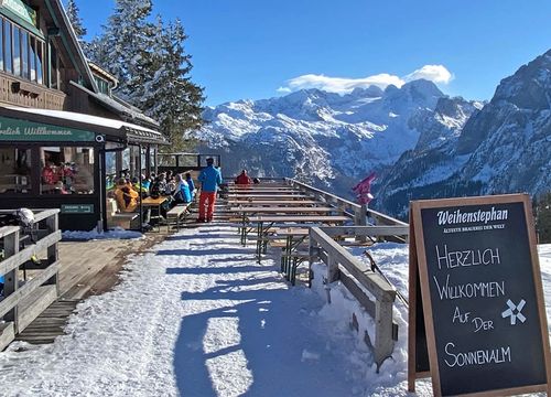 Verschneiter Bergblick mit einer Almhütte und Sitzgelegenheiten im Freien. Auf einem Schild steht „Herzlich Willkommen auf der Sonnenalm“. Die Menschen genießen die Landschaft.