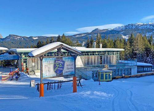 Bergstation eines Sessellifts, mit angebautem modernen Restaurant, umgeben von Bäumen und Bergen unter einem klaren blauen Himmel.