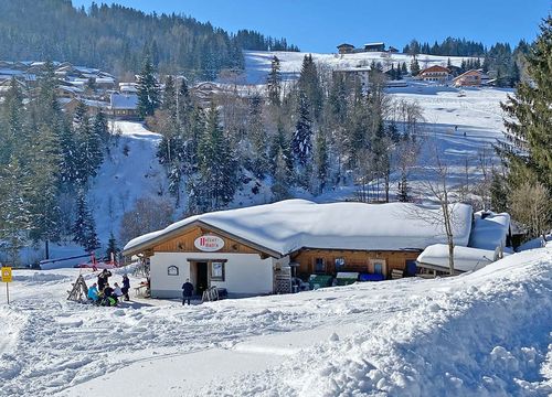 Eine schneebedeckte Hütte in einer Berglandschaft, vor der sich Menschen versammelt haben. Unter einem klaren blauen Himmel sind Tannenbäume und Skipisten zu sehen.