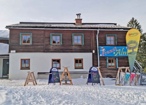 An alpine wooden building with colourful window frames, surrounded by snow. There are wooden ski and snowboard stands and advertising banners in front of the hut.