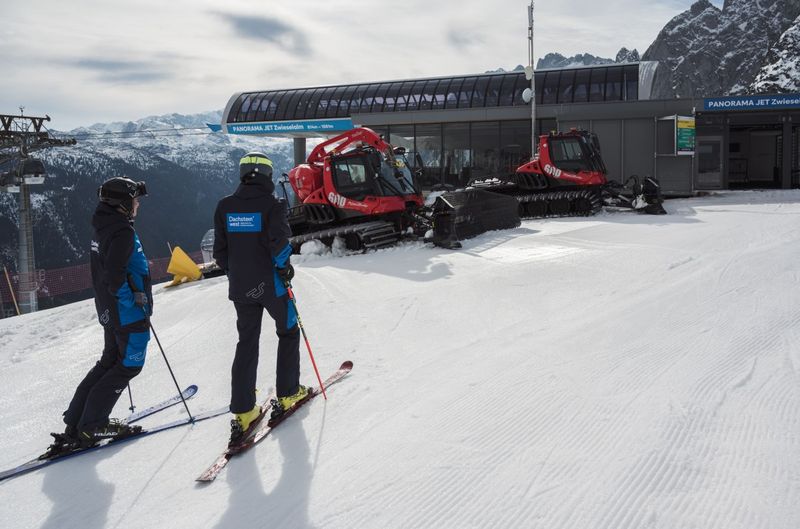 Two skiers stand on a snow-covered piste near a ski lift station, with red snow groomers and mountain peaks in the background.