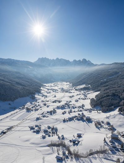 Luftaufnahme eines schneebedeckten Tals mit verstreuten Häusern, umgeben von Bergen unter strahlender Sonne und blauem Himmel.