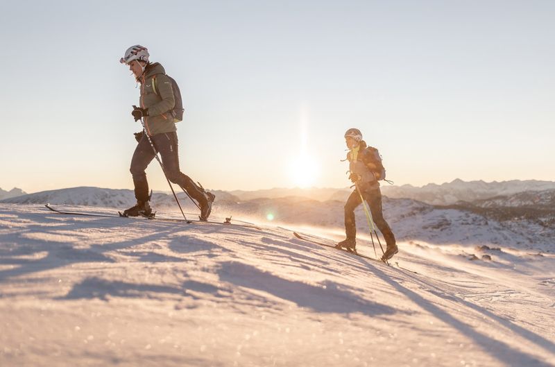Zwei Skifahrer steigen bei Sonnenaufgang einen verschneiten Hang hinauf, mit Bergen im Hintergrund und der Sonne, die ein warmes Licht auf sie wirft.