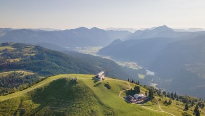 Luftaufnahme einer üppig begrünten Hügelkuppe mit einem Seilbahngebäude und einer Almhütte, umgeben von Bergen und Tälern unter einem klaren blauen Himmel.