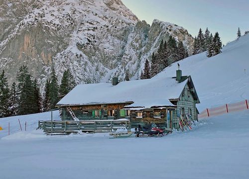 Eine schneebedeckte Hütte mit einer Holzterrasse liegt in einer Gebirgslandschaft, umgeben von Bäumen und Skiausrüstung.