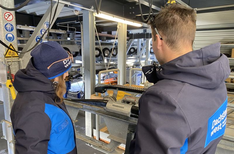 Zwei Personen in blauen Jacken arbeiten in einer industriellen Umgebung an Maschinen, wobei sie sich auf ein Metallteil konzentrieren. Im Hintergrund sind Regale mit Geräten zu sehen.