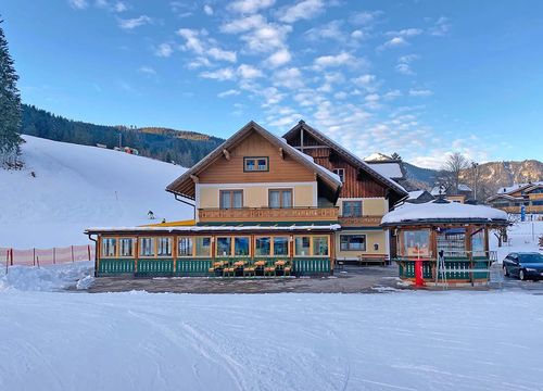 A cosy ski hut with a snow-covered roof, surrounded by snow-covered slopes and evergreen trees under a clear blue sky.