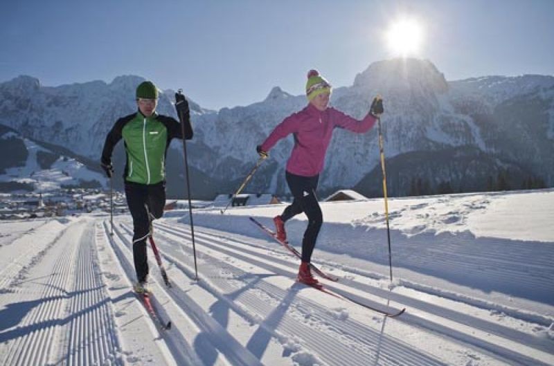 Zwei Personen beim Skilanglauf an einem sonnigen Tag, mit verschneiten Bergen im Hintergrund.