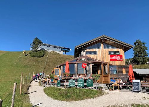 A mountain hut with outdoor seating on a sunny day, surrounded by a grassy hill and blue skies.