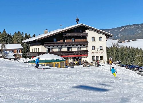 Skifahrer, die einen verschneiten Hang in der Nähe eines großen Hotels unter blauem Himmel hinunterfahren, mit bewaldeten Hügeln im Hintergrund.
