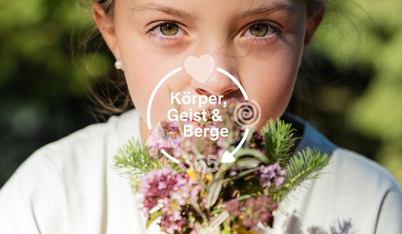 Ein Mädchen hält einen Strauß Wildblumen neben ihrem Gesicht. Der überlagerte Text oder das Logo lautet „Body, Mind & Mountains“ mit den Symbolen „Herz“, ‚Spirale‘ und „Berg“.