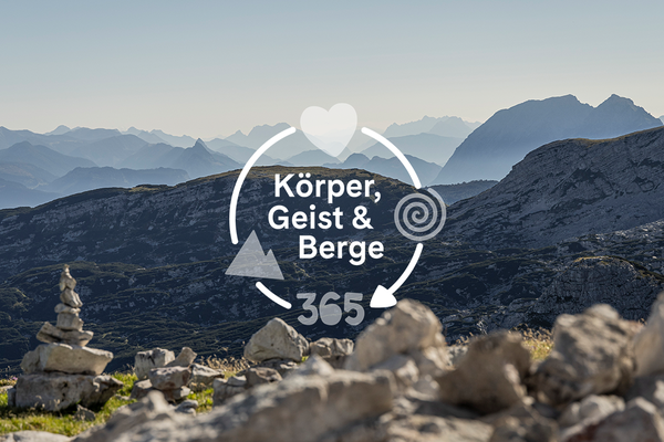 Steinmandln im Vordergrund, dahinter das karge Dachsteinplateau mit weichem Morgenlicht über den Bergketten mit Logo "Körper, Geist und Berge" darüber
