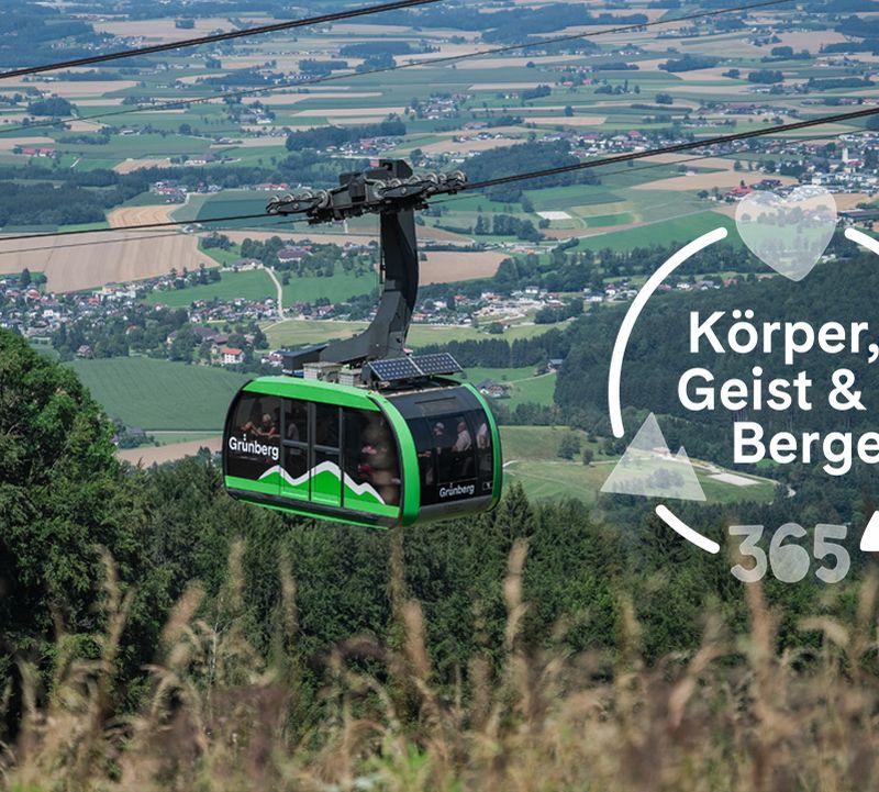 Grüne Gondel der Grünbergbahn vor weitem Talblick mit Feldern und Orten. Overlay: Körper, Geist & Berge 365.