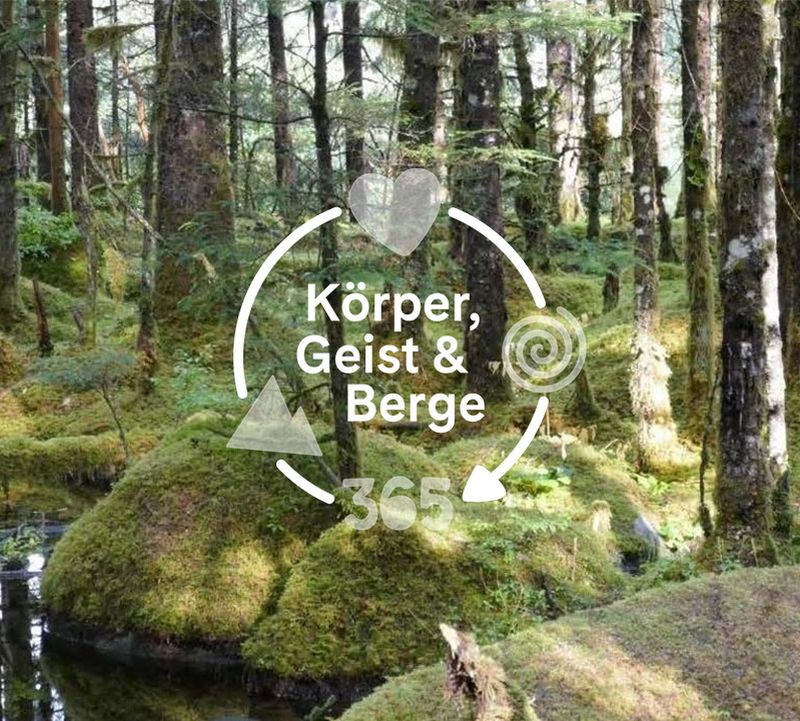 Ein üppiger, moosbewachsener Wald mit einem kleinen Bach. Der überlagerte Text bzw. das Logo lautet „Körper, Geist & Berge“ mit den Symbolen "Herz", "Spirale" und "Berg".